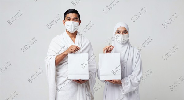 The image shows a man and a woman wearing white Ihram clothing, with face masks on, indicating they are preparing for Hajj or Umrah. Each is holding a white bag, standing against a plain white background.