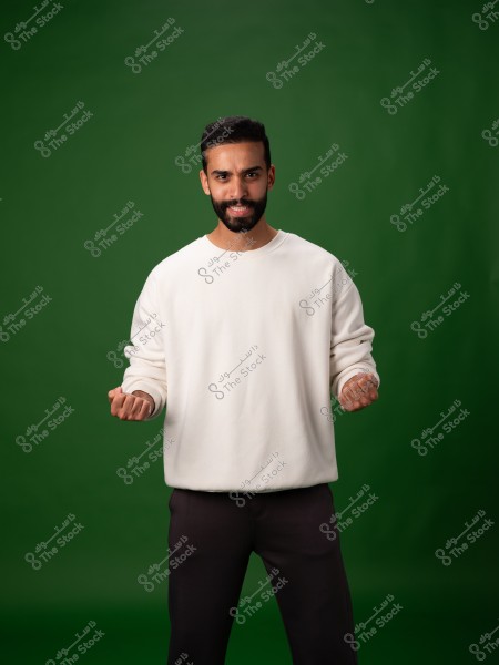 A portrait of a man with a beard and short hair wearing a white sweater and dark pants. He stands in front of a green background.