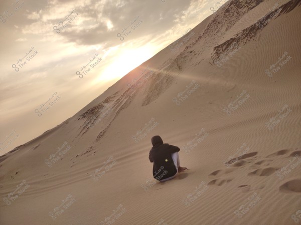 Image of a person sitting on sand dunes in the desert at sunset. Footprints can be seen in the sand while the sun\'s rays create a warm orange glow across the sky. The person is dressed in dark clothing, viewed from behind, and appears to be enjoying the tranquil natural scenery.