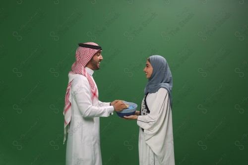 امرأة سعودية ترتدي العباءة و رجل سعودي يرتدي الثوب و الشماغ يتبادلان الهدايا وهما مبتسمان و سعيدان احتفالًا بالعيد