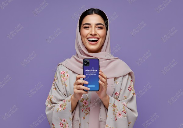 A portrait of a smiling woman holding a smartphone displaying the \"AlimaPay\" logo. She is wearing a pink hijab and an abaya adorned with floral patterns. The background is light purple. The clothing style appears traditional and may suggest a Middle Eastern origin.