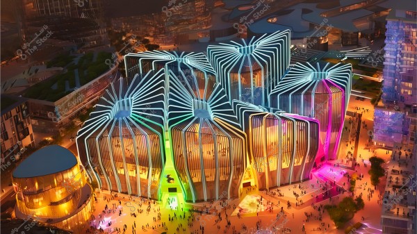 A nighttime view of a modern futuristic building, illuminated with bright colors such as purple, blue, and yellow. The building features distinctive geometric designs resembling flowers or decorative webs. People are seen walking around the structure in the open space.