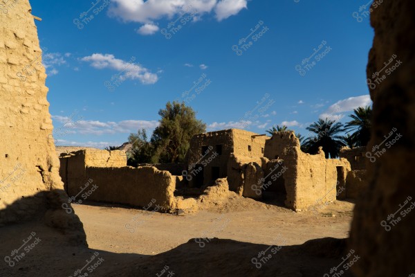 تظهر الصورة منظرًا لمبانٍ طينية قديمة تقع تحت سماء صافية زرقاء مزيّنة ببضع غيوم. تبدو الجدران الطينية بنيّة اللون ومتآكلة قليلاً, وتحيط بمساحة مفتوحة واسعة. تظهر أشجار نخيل خضراء كثيفة في الخلفية, مما يوحي بمكان صحراوي تاريخي.