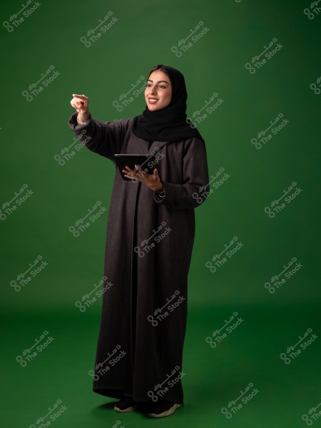 The image shows a woman wearing a black abaya and black hijab, holding a tablet in her left hand and pointing forward with her right hand. The background is green.