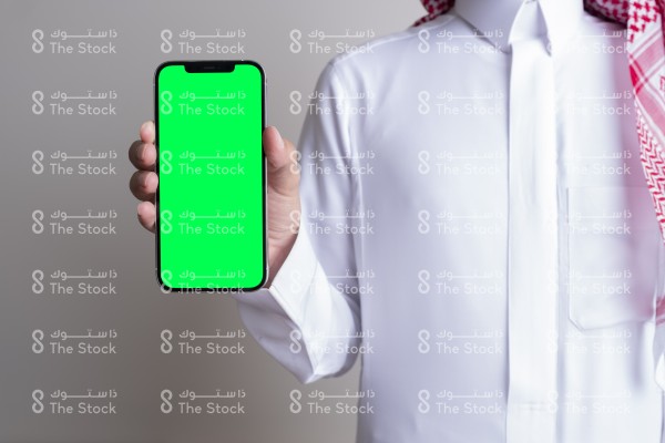 صورة ملتقطة بهاتف لرجل سعودي يرتدي الزي السعودي التقليدي يمسك بالجوال ويعرض شاشته بوضوح