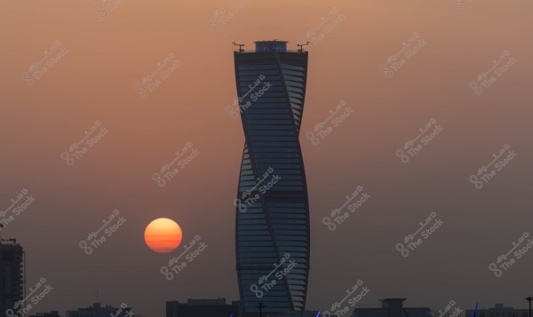 An image of an architecturally designed skyscraper with a slight twist in the middle, located in an unspecified city, with the sun setting in the background, giving the sky a warm orange hue. There are other buildings in the foreground.