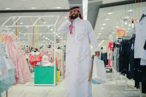 رجل سعودي يرتدي الزي السعودي التقليدي يحمل أكياس التسوق بينما يقوم بإجراء مكالمة هاتفية