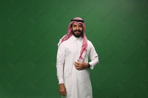 رجل سعودي يقف على خلفية خضراء يرتدي الثوب و الشماغ، يقوم بمسك الشماغ وهو مبتسم ، احتفالات العيد