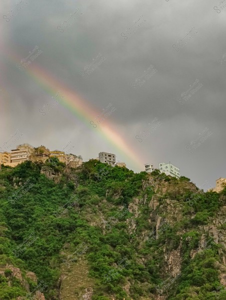 تظهر في الصورة قوس قزح يمتد عبر سماء ملبدة بالغيوم فوق تلة خضراء وكثيفة بالنباتات. تُرى على قمم التلة عدة مبانٍ بألوان فاتحة, بعضها بلون البيج والآخر باللون الرمادي. الألوان المختلفة لقوس قزح تضيف لمسة من الجمال إلى المشهد الطبيعي.