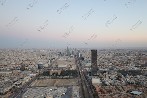 Aerial view of Riyadh city in Saudi Arabia showcasing tall buildings and the central road. The Kingdom Tower is prominently in the center, surrounded by numerous residential and commercial buildings. The sky is clear with light clouds in the background.