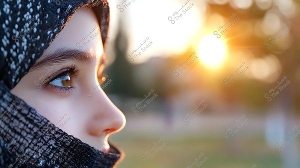 A side profile portrait of a young girl wearing a patterned black hijab, gazing towards the bright sun in the background. The golden light from the sun emits a warm glow. The background is blurred, highlighting the facial features and details of the hijab.