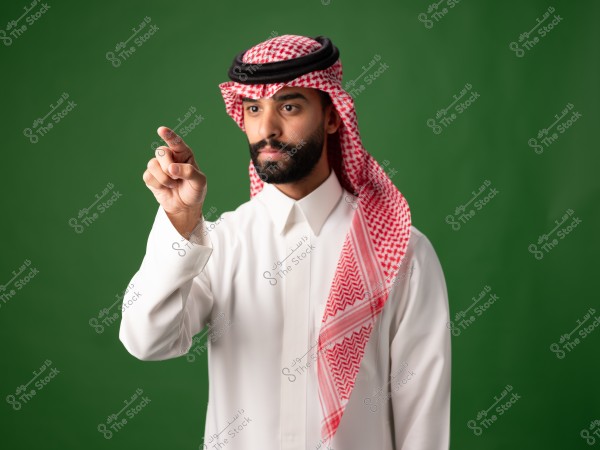Image of a man wearing traditional Saudi attire, consisting of a white thobe and a red and white ghutra with a black agal. The background is green, and the man is extending one hand forward, pointing at something.