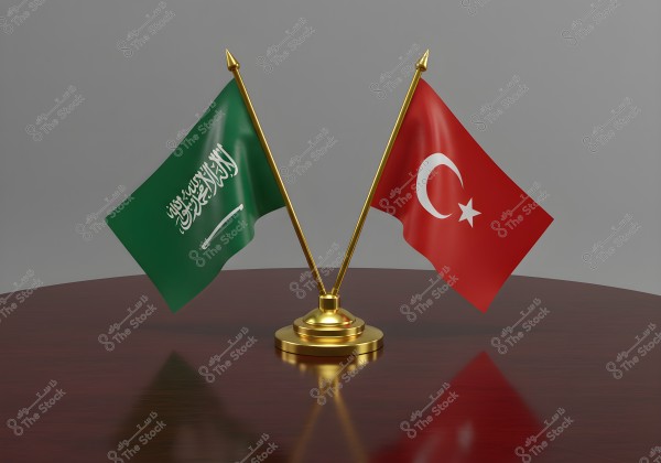An image of two national flags, the Saudi flag with a green background and white Arabic inscription with a white sword, next to the Turkish flag with a red background and a white star and crescent on golden crossed poles atop a wooden table.