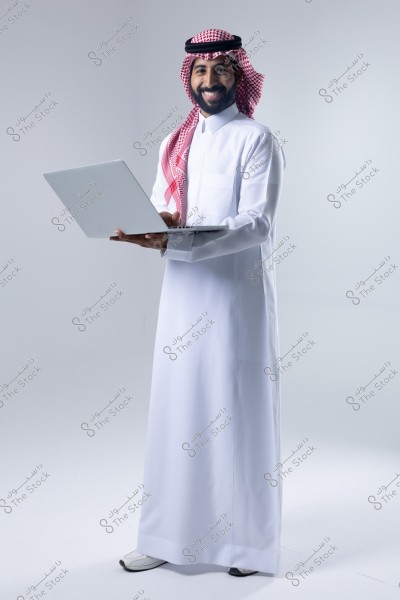 An image of a man wearing traditional white thobe and a red checkered shemagh with an agal, holding a laptop and smiling. The backdrop is white, suggesting the attire might be Saudi in origin.