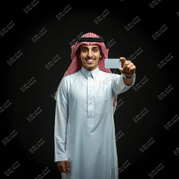 A portrait of a man wearing traditional Saudi attire, a white thobe, along with a red and white checkered shemagh and black agal. He is smiling and holding a white card in his right hand. The background is black.