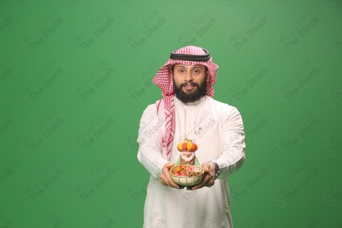 شاب سعودي على خلفية خضراء يرتدي الثوب و الشماغ، يقوم بعمل حركات بيده،يمسك بسلة الحلوى و يقدمها، احتفالات العيد