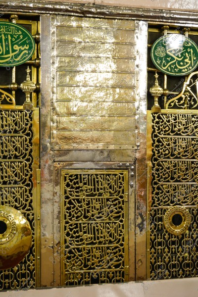Image of a section of the golden grille of the Prophet Muhammad’s tomb in Al-Masjid an-Nabawi, Madinah, Saudi Arabia. The intricate design features engraved Arabic script and decorative inscriptions. The golden frame has complex engravings and is adorned with green circles with white script.