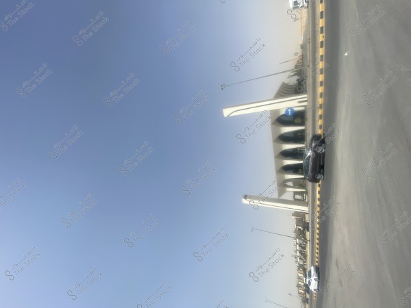 منظر لواجهة مبنى حديث مع أعمدة مميزة وشكل معماري فريد, يظهر خلفها السماء الصافية. في المقدمة يوجد طريق مع علامتين للمرور وسيارتين متوقفتين, واحدة باللون الأبيض وأخرى باللون الأسود, بجانب حديقة صغيرة.