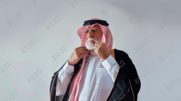 A portrait of an elderly man wearing traditional Saudi attire, including a red and white checked headscarf and a black cord. He is adjusting his garment at the collar. The man has a white beard and is wearing a black cloak with gold embroidery. The background is a simple light color.