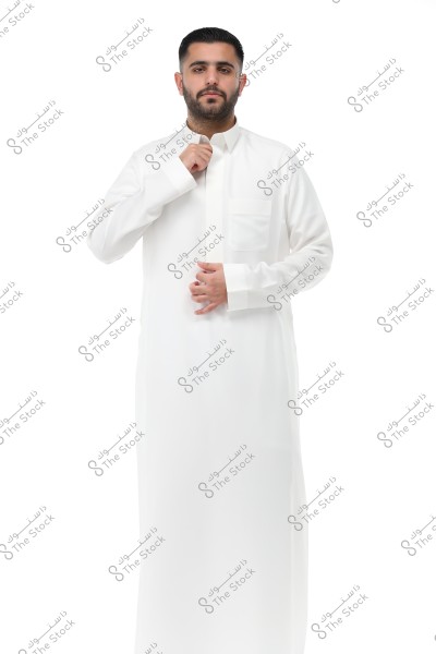 Portrait of a man wearing a traditional white thobe, possibly from Saudi Arabia. The man stands against a white background, adjusting the collar of his thobe with his right hand. He has a serious expression, trimmed beard, and short hair.