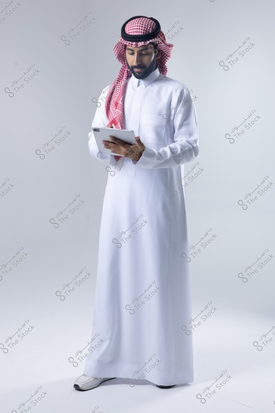 A portrait of a man wearing traditional Saudi attire, featuring a long white thobe with a red and white checkered headscarf and a black agal. He appears to be using a tablet. The photo is taken in a studio with a white background.