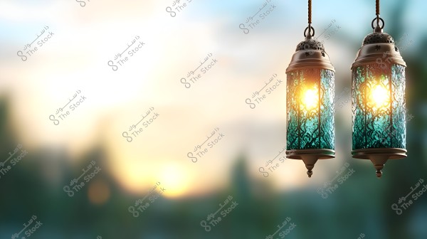مصباحان تقليديان معلقان يتألقان بالضوء الذهبي في وسط الصورة. المصابيح مصنوعة من زجاج مزخرف بلون أزرق وأخضر ومحيطة بإطارات معدنية. الخلفية غير واضحة تُظهر غروب الشمس بألوان دافئة, مع تدرج من الأزرق إلى الأصفر فوق منظر طبيعي غير واضح.