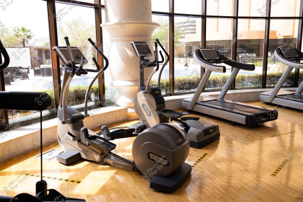The image shows an indoor gym with various fitness equipment, including treadmills and stationary bikes. The floor is polished wood, and large windows offer a view of the outdoors with trees and a swimming pool. Natural light fills the area through the wide windows, giving a fresh and comfortable atmosphere.