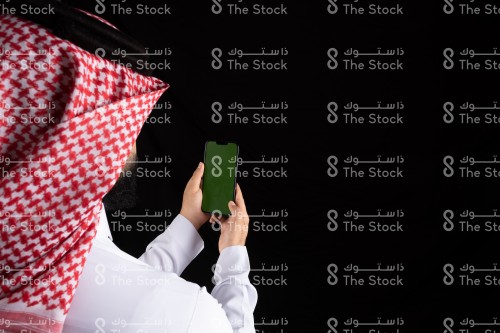 رجل سعودي يرتدي الثوب يحمل الجوال بيده وهو ينظر إليه مع خلفية سوداء، رجل ينظر الى شيء على الجوال