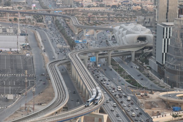 Aerial view of a modern city featuring a network of highways crowded with vehicles. The image shows a metro train traveling on an elevated track, alongside tall buildings and a uniquely designed modern structure resembling waves. The scene reflects urban development and heavy traffic activity in the city.