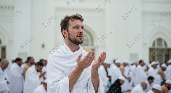 The image shows a group of people performing the Hajj or Umrah rituals. The person in the foreground is wearing a white ihram and standing with his hands raised in prayer, while others gather in the background wearing white ihram garments. It appears to be one of the holy sites in Mecca, surrounded by white walls.