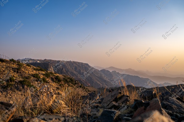 A natural scene featuring towering mountains under a clear blue sky with a faint sunlight illuminating the horizon. In the foreground, there are rocks and small dry plants, while the mountains stretch into the background with beautiful light gradients reflecting from the sun.