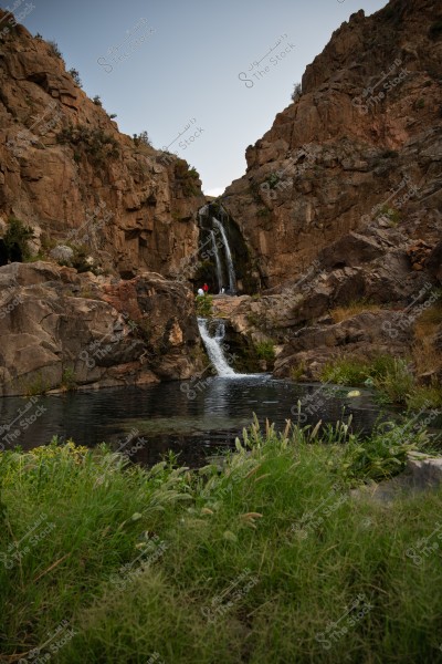 An image showing a beautiful waterfall cascading between high rocks in a valley surrounded by massive rock formations. The water flows into a small pool below, bordered by green plants along its banks. In the background, a clear blue sky is visible.