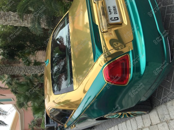 A luxury car with gold and green colors parked on a brick pavement. The license plate indicates Dubai. Palm trees are visible in the background with a pink building on the left side.