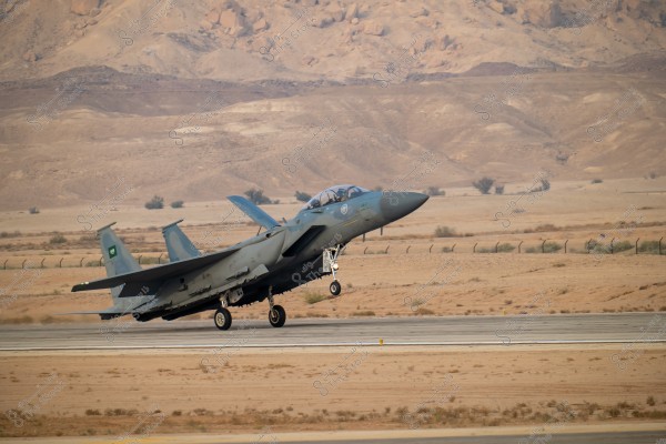 A fighter jet taking off, featuring the Saudi Arabian insignia, departing from a runway in a desert environment with rocky mountains in the background. The sky is clear and the weather appears sunny.