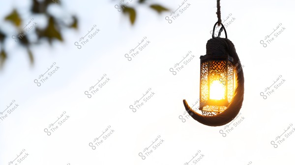 A decorative metal lantern hanging on a rope, illuminated against a bright outdoor scene. The lantern is suspended from a rope, glowing with a warm light, and some tree leaves are blurred in the top left corner of the image.