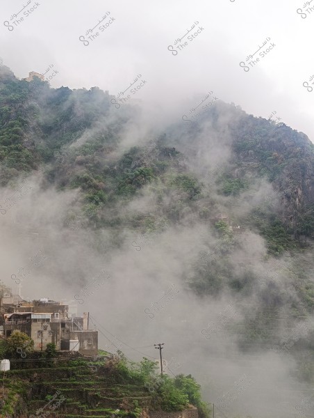 The image shows mountains covered in thick mist in a green area, with houses visible at the bottom of the mountain displaying clear details of rooftops and electricity poles in the foreground. The surrounding rocks and foliage add a natural and majestic atmosphere to the scene.