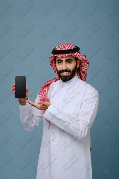 بورتريه شاب سعودي يرتدي الزي السعودي يشير إلى هاتفه ويتفاعل معه، وتظهر على ملامحه السعادة ، على خلفية سماويه