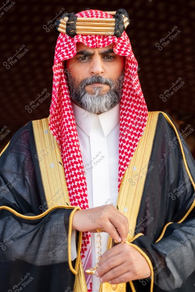 صورة لرجل يرتدي زيًا تقليديًا سعوديًا, يشمل ثوبًا أبيض وعباءة سوداء مزينة بخطوط ذهبية. يغطي رأسه شماغ أحمر وأبيض مع العقال الأسود الذهبي. الرجل يمسك بمسبحة في يده ويقف أمام خلفية داكنة.