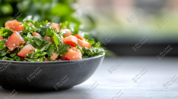 An image of a bowl containing fresh tabbouleh salad made with chopped parsley and diced tomatoes. The salad is served in a dark-colored bowl placed on a gray surface, with a blurred light green background.