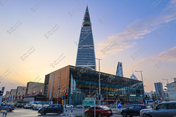 The image shows Al Faisaliah Tower in Riyadh, Saudi Arabia, featuring its distinctive pyramid-shaped design and golden spherical cap. In the foreground, there is a shopping center with famous brand names like \"Gucci,\" \"Cartier,\" and \"Burberry.\" Cars are gathered at a traffic light, indicating a busy street. The sky is mostly clear with a few scattered clouds at sunset.