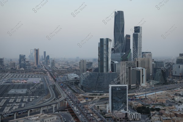 The image shows a view of Riyadh city in Saudi Arabia, featuring prominent skyscrapers like the Kingdom Tower and other modern buildings. A busy highway with cars flows under overpasses, with the sky in the background appearing pale during sunset.