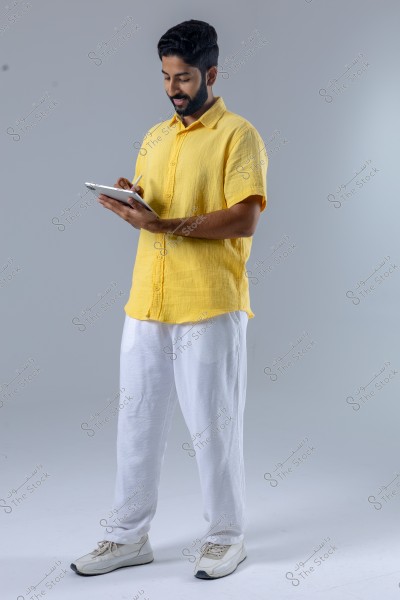 The image shows a man standing, wearing a yellow shirt and white pants with white sneakers. He holds a tablet and is using a stylus to write on it. The background is a simple light gray, and the man appears focused on his activity.