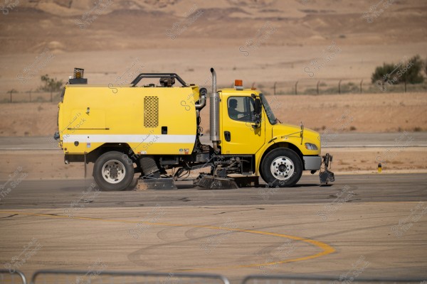 A small yellow truck used for cleaning asphalt at an airport. The truck has brushes at the front and is moving on the airport runway with a desert landscape in the background.