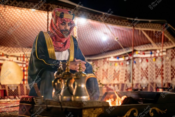 صورة لرجل يجلس في خيمة تقليدية ويبدو أنه من شبه الجزيرة العربية. يرتدي الزي التقليدي الذي يشمل الثوب الأبيض والعباءة السوداء المزينة بالذهبي والغترة الحمراء والبيضاء مع العقال الأسود. أمامه جلسة تقليدية تحتوي على القهوة وتوجد عدة أباريق نحاسية على النار المشتعلة. الخيمة تحتوي على سجادات وأنوار ملونة تضيف جوًا دافئًا للمكان.
