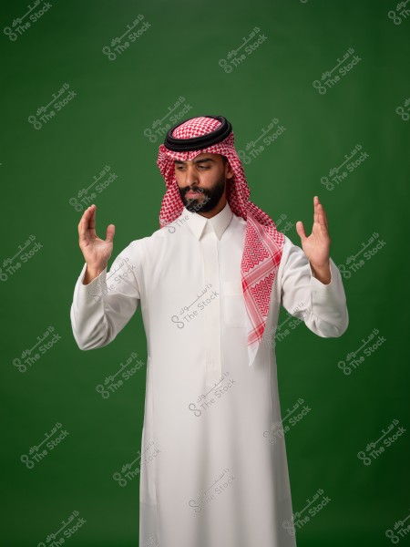 ** A man standing wearing a white thobe and a red and white ghutra with a black agal. He appears to be explaining or speaking, with his hands raised. The background is green.\r\n\r\n**