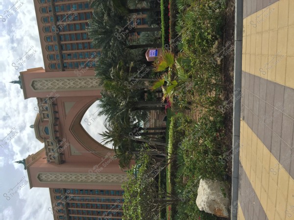 The image shows part of a grand hotel featuring a large central arch and a facade with geometric designs. The front garden of the hotel is surrounded by lush tropical plants and trees. The sky is cloudy, adding a natural and serene ambiance.