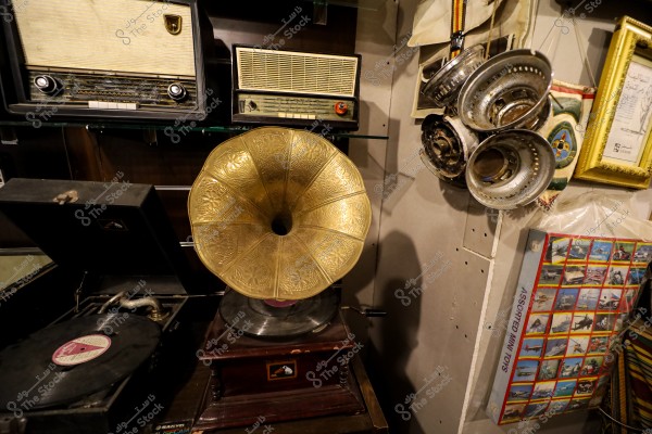 An image showing a collection of vintage items, including a golden phonograph with engravings displayed on a wooden base. Next to it is an antique radio. Hanging on the wall are metal objects and several framed pictures and photos.