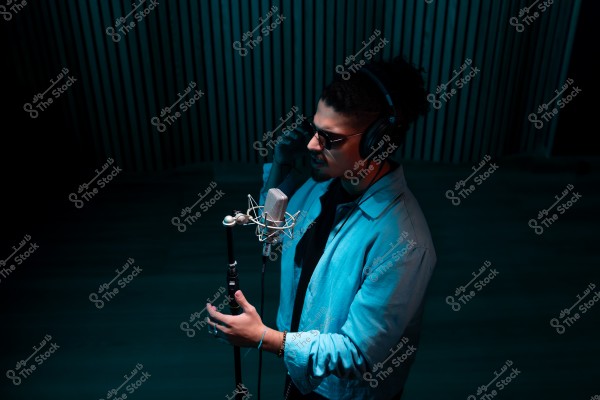 رجل يرتدي سماعات رأس ونظارات شمسية يقف أمام ميكروفون في استوديو تسجيل مضاء بإضاءة زرقاء.