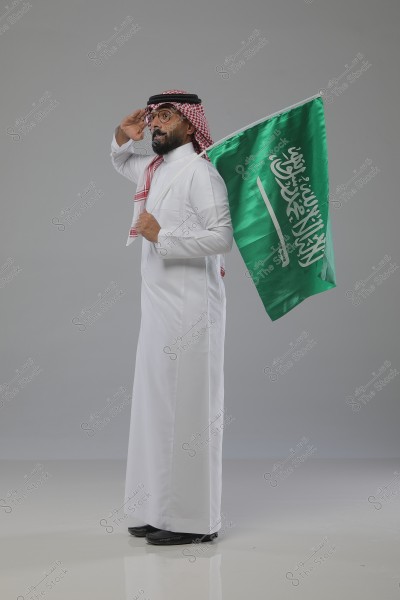 An image of a man wearing a white thobe standing next to the flag of Saudi Arabia. The man is wearing a traditional headscarf and agal and is saluting. The gray background highlights the vibrant colors of the Saudi flag.