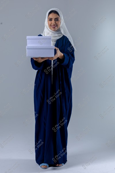 Image of a woman wearing a dark blue abaya and a white hijab, holding two white boxes. She is standing against a gray background and smiling at the camera. The clothing style may suggest Saudi Arabian culture.